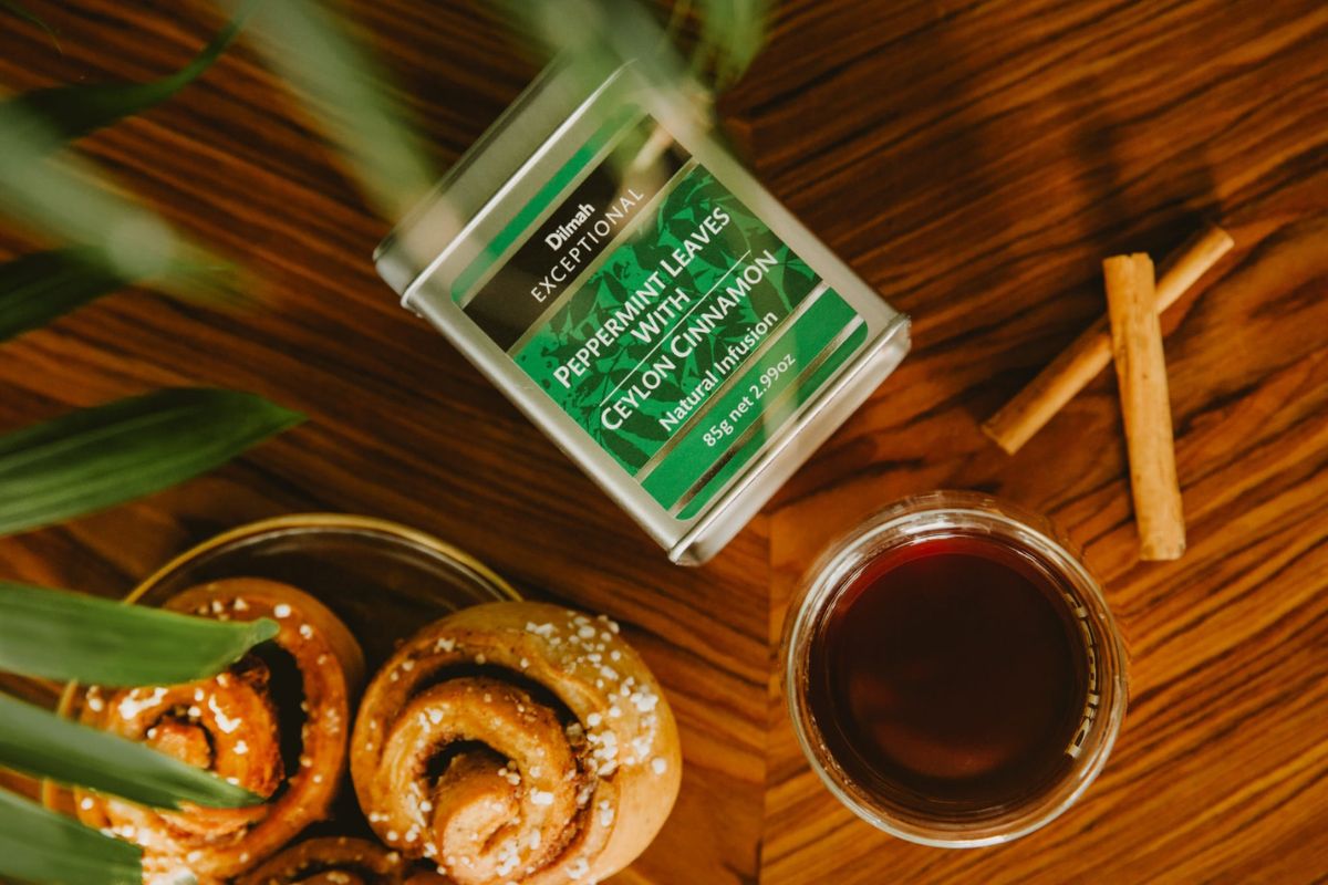 Napar z mięty z cynamonem w przezroczystej szklance. Obok bułeczki cynamonowe, laski cynamonu i opakowanie Dilmah Exceptional Peppermint Leaves with Ceylon Cinnamon.